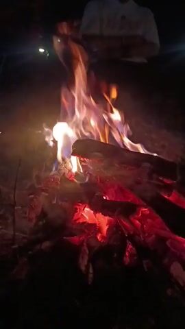 The video shows a campfire burning at night.  The flames are a mix of red, orange, and white, licking around logs arranged in a pile.  A person stands behind the fire, their face obscured by darkness.  A few faint lights appear in the background, adding to the night ambiance.  The fire emits smoke and ash, adding a dynamic element to the scene.