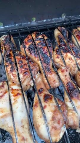 The video shows chicken drumsticks cooking on a charcoal grill. The camera is angled downwards, focusing on the grill's surface and the chicken. The drumsticks are arranged on the metal grates of the grill. Some have visible grill marks. Underneath the grates are hot coals, which produce some smoke. The camera slightly moves and adjust position. Some green shrubbery is visible in the background.