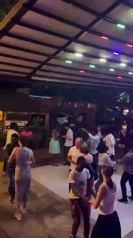 The video shows a group of people dancing and socializing outside a restaurant or bar at night. Some people are dancing in pairs on a tiled area under a covered roof with string lights. Other people are standing around talking in smaller groups, some near the entrance of a building. The environment includes plants, and the architecture appears to have a tropical or resort style. The lighting is dim and colorful, creating a festive atmosphere.