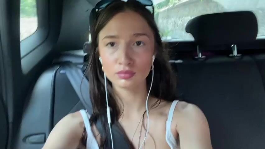 This media file shows a young woman sitting in the passenger seat of a car. She is wearing a white tank top, a black belt, and a floral skirt. She has long dark hair and is wearing white earbuds. She appears to be looking at the camera with a neutral expression. The car appears to be moving, as suggested by the blurred background. The interior of the car is dark, with black leather seats. A seatbelt is visible across the woman's chest. In the background, there are hints of trees and foliage, suggesting an outdoor setting. The overall impression is that the woman is on a car journey. There is no visible text in the media file.