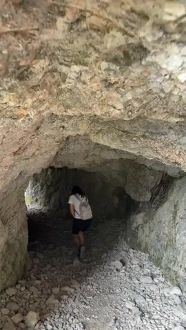 The video shows a person walking through a rocky tunnel. The person is wearing a white t-shirt, black shorts, and a backpack. The tunnel walls are made of rough, light-colored rock. The ground is covered in small rocks and pebbles. The tunnel appears to be natural, or at least very old and unmaintained. The lighting is dim inside the tunnel, but the end of the tunnel is brightly lit, suggesting an exit into an outdoor area. The camera is moving, following the person through the tunnel.