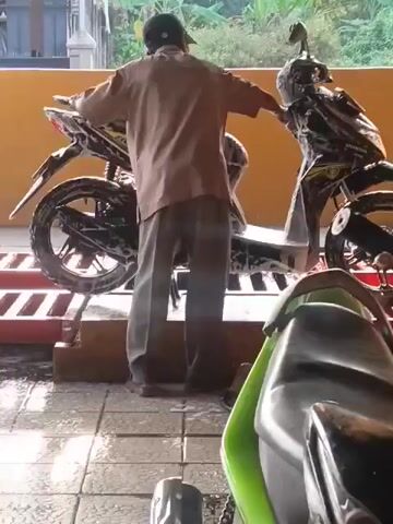 The video shows a person washing a black and yellow motorcycle. The person is wearing a brown shirt and gray pants and has a cap on. The motorcycle is covered in soap suds. The person is using a sponge or cloth to wash the motorcycle. There is another green motorcycle in the foreground. The scene appears to be outdoors with a yellow wall in the background. The ground is wet and soapy, suggesting it is a car wash or a similar area.