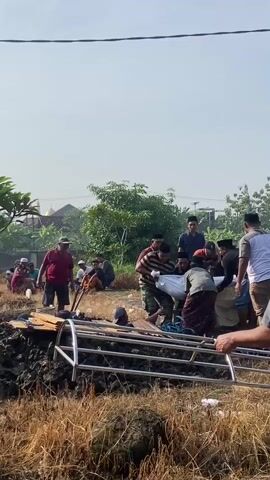 The video shows a funeral scene. A group of men, some wearing Muslim caps, are lowering a white-shrouded body into a grave. The grave is located in a grassy area with a mosque visible in the background. Some people are sitting in the background observing the burial. A silver frame is partially visible in the foreground. Overhead power lines are strung across the sky. The sky is overcast, but it appears to be a bright day.