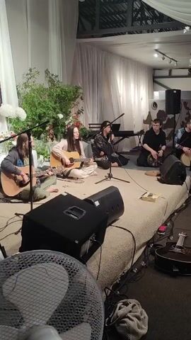 A group of six musicians performs on a raised platform covered with a textured rug. The platform is illuminated by soft lighting and adorned with white floral arrangements and greenery. In the foreground, a large black speaker is positioned on the floor, along with a white fan. To the right, an acoustic guitar rests on the floor. On the platform, from left to right, a man with long hair plays an acoustic guitar. Next to him, a woman sings and plays an acoustic guitar. To her right, a person with dark hair sings into a microphone. Further back and to the right, three men are seated. One man plays a hand drum, another plays a keyboard, and the third man also plays a hand drum. The musicians are dressed casually. The background features sheer white curtains and a glimpse of a tent-like ceiling structure. A microphone is positioned in front of the singing woman.