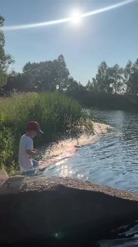 A young person wearing a red baseball cap and a white t-shirt kneels on a concrete edge next to a body of water. The sun is bright in the sky, casting a glare on the water's surface. Tall green reeds grow along the bank of the water. Trees form a treeline in the background. The person is reaching out towards the water with their hands.