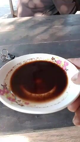 The video shows a close-up of a white bowl filled with a dark liquid, possibly coffee. The bowl has a floral pattern around its rim. A set of keys with a silver ring is visible to the left of the bowl. The bowl rests on a weathered wooden surface, suggesting an outdoor setting. The lighting indicates it is daytime, with shadows cast on the surface. In the background, a person is partially visible, seated and out of focus. The liquid in the bowl appears to be disturbed by a swirling motion.