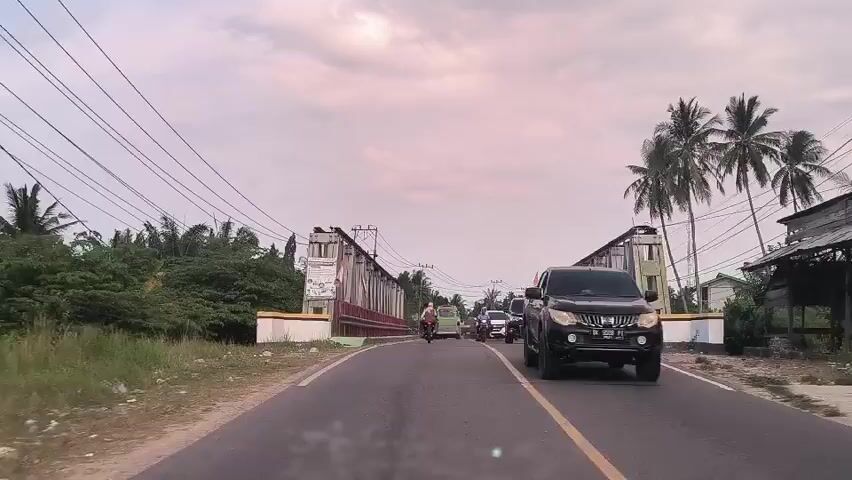 A vehicle-mounted camera travels down a road toward a bridge. The bridge is a metal truss bridge with a red and yellow handrail. The road has a yellow dividing line. Other vehicles, including cars and motorcycles, are on the bridge. There are palm trees and power lines visible along the sides of the road. The sky is overcast.