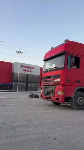 The video shows a red semi-truck with a large white cylindrical trailer. The truck is on a dirt road next to a building that says "FASHION MALL" in large letters. The truck appears to be moving or has just stopped. A man can be seen in the driver's seat. The trailer has red markings on its side. The sky is clear and appears to be late afternoon or early evening.