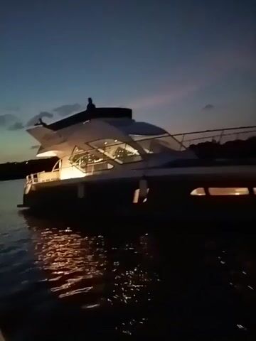 The video shows a large, modern yacht illuminated with lights, at dusk or night. The yacht is docked or anchored in calm water, with reflections of the lights visible on the water's surface. The sky is a dark blue with hints of pink from the setting sun. A person appears to be standing or sitting on the upper deck of the yacht. Some trees or vegetation can be seen on the distant shoreline. The camera appears to be on a smaller boat or dock, providing a side view of the yacht.  The video is somewhat shaky.