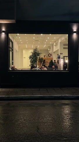 The video captures an exterior view of a building through a large, rectangular window at night. The building's facade is dark, with vertically aligned panels and symmetrical lighting fixtures flanking the window. The street outside is wet, reflecting the ambient light. Inside the brightly lit building, several people are visible in a waiting area, seated on what appears to be a large sofa. There is a potted tree nearby, and various objects and furniture pieces are scattered throughout the room. One person is standing and appears to be occupied with something near a column. The focus remains on the interior scene, showcasing a well-lit, occupied space viewed from a dark, wet exterior.