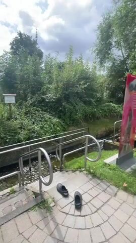 A person wearing a red bandana and a dark jacket is walking through a shallow stream. They are wearing shorts and appear to be barefoot. There are metal railings on either side of the stream for support. The stream is surrounded by lush green vegetation and trees. The ground in front of the stream is paved with bricks. In the background, there is a grassy field and more trees. The sky is partly cloudy.