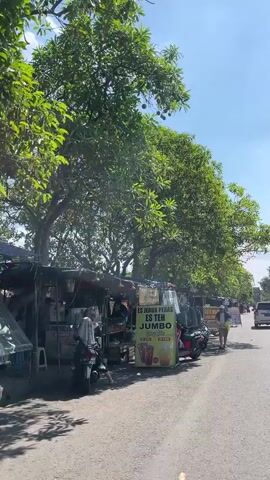 A street scene in Sumokali, Indonesia, under a clear blue sky. The sun is shining brightly, casting shadows and illuminating the lush green trees that line the road. The setting appears to be a roadside market or collection of vendors. Several small stalls are set up, selling drinks and possibly other goods.

In the foreground, a red scooter is parked, and next to it is a prominent sign advertising "ES JERUK PERAS" and "ES TEH JUMBO" (freshly squeezed orange juice and jumbo iced tea). The price "RP 5.000" is also visible. Several people are interacting with the stalls or walking along the street. One person is seated on a motorcycle, and others are standing or moving about.

Behind the stalls, more trees and a glimpse of other structures can be seen. The overall atmosphere suggests a warm, bustling day in a tropical or subtropical region. The presence of multiple scooters and the style of the stalls indicate a common mode of transport and local commerce in the area. The scene is vibrant and captures the essence of daily life on a sunny day in Indonesia.