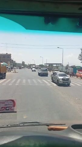 The video, filmed from inside a vehicle with a green-tinted windshield, shows a street scene during a sunny day.  The vehicle is positioned at a crosswalk on a multi-lane road, where vehicles including a truck and a pickup truck are visible. Buildings and street lights line the sides of the road. A few pedestrians are walking along the sidewalks. The camera remains stationary throughout the video.