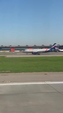 From an airplane window, a tarmac is seen during daylight. The airport terminal is in the background. Several airplanes with blue, white, and red markings are lined up at the terminal. An air traffic control tower stands nearby. One airplane crosses the tarmac as seen through the window.