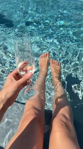 This media file shows a close-up view of a person's legs submerged in a swimming pool, with one hand holding a champagne flute filled with a bubbly beverage. The water is clear and blue, with sunlight creating dappled patterns on the surface and the submerged legs. The person's bare feet are visible in the water, and their legs appear tanned. The focus is on the sensory experience of relaxation and enjoyment, suggesting a leisurely moment. The setting appears to be a sunny outdoor location, possibly during the daytime, given the bright light and clear water. The overall mood is one of leisure and indulgence. There is no visible text in the media file. The location context is Asciano, Italy.