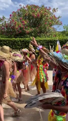 A group of people in colorful Hawaiian shirts and grass skirts are participating in a parade. They are wearing straw hats decorated with feathers and flowers. Some are holding flags, including a rainbow flag and a flag with transgender colors. The people are smiling and appear to be enjoying themselves. The background shows a lush green hedge and a tree with pink flowers. The ground is paved with light-colored stones. The sky is blue with some clouds. One person in the foreground is holding a fan made of what appears to be money.