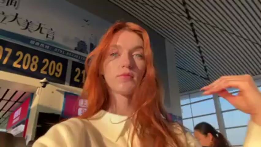 A woman with long red hair and light eyes stands indoors in front of a large sign with numbers on it. She is wearing a light-colored collared shirt. She initially gestures with her hand in front of her face and then moves her hand to her hair behind her ear. In the background, there is a partially visible woman and a ceiling with dark lines. The lighting is bright, seemingly from sunlight.