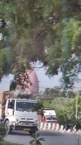 A video taken from a moving vehicle shows a large, pink, petal-shaped structure in the background. The structure appears to be a building or monument. Trees and foliage partially obscure the view. In the foreground, a portion of a white truck with a tarp-covered cargo bed is visible on the right side of the frame, suggesting the video was filmed from a roadway. A utility pole stands prominently in the middle of the frame. The sky is overcast. The camera is in motion, causing a slight blur in the background and foreground elements.