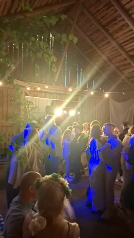 The video shows a crowd of people dancing inside a wooden barn. The lighting is dynamic, with changing colors from blue to red to green, illuminating the faces and bodies of the dancers. Some people are dancing in pairs, while others are standing in groups. The ceiling of the barn is made of exposed wooden beams, and there are string lights hanging across the space. The atmosphere appears festive and celebratory.