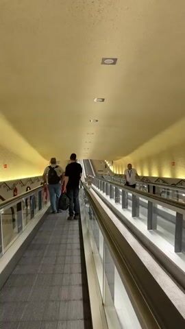 The video shows a long moving walkway in a seemingly public space, possibly an airport or train station, with a high, curved ceiling. Two individuals walk away from the camera on the left-hand side of the walkway, carrying bags. On the right-hand side, another individual walks towards the camera. The walkway is illuminated by warm lighting, creating a yellow hue on the ceiling and walls. Metal railings with glass panels separate the walkways from each other and the sides. There are some square light fixtures on the ceiling. The scene appears to be in motion, suggesting the walkway is operational.