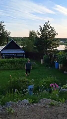 A man is in his backyard, which has a lawn, a fence, a house, and trees. He is playing with a boy and they appear to be throwing something. The sky is blue with some clouds. The man is wearing a dark t-shirt and shorts. The boy is wearing a hoodie and shorts. There is a yellow object in the air.