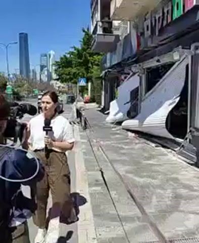 The video shows a woman holding a microphone standing on a sidewalk, apparently a reporter giving a news report. To the reporter’s left is a cameraman operating a camera. A little boy is walking behind the reporter. Buildings and streets are in the background with a very tall building further in the distance. The storefront of the building near the reporter appears to be damaged, with white metal siding bent and dislodged. The sidewalk is made of gray concrete.