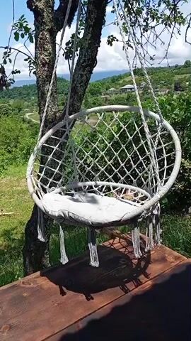 The video shows a hanging chair suspended from a tree. The chair is made of white rope in a net pattern with tassels hanging from the bottom. A white cushion is inside the chair. The chair is positioned on a wooden platform. In the background, there is a view of rolling green hills and some buildings. The weather appears to be sunny. The camera is positioned slightly below the chair, looking up.
