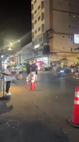 The video shows a nighttime street scene in what appears to be a tourist area. The street is lined with buildings, some with signs in English. There are people walking, some crossing the street, and others standing near food stalls. Several vehicles are visible, including cars, motorcycles, and a small red truck. Orange traffic cones are placed along the street, possibly indicating a construction zone or traffic diversion. The scene is lit by streetlights and the lights from businesses. The overall impression is a busy, lively street at night.