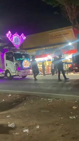 At night, a white truck with colorful neon lights is parked on the side of a road. People are seen near the truck, some appearing to be interacting with it. In the background, a yellow building with the Shell logo is visible, suggesting a gas station or store. A motorcycle with two riders passes by in the foreground. The ground is dirt and littered with trash.
