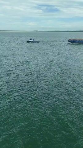 The video shows a tugboat towing a large barge on a body of water. The water is choppy, and the sky is overcast. In the distance, there are other boats visible. The scene appears to be a maritime environment.