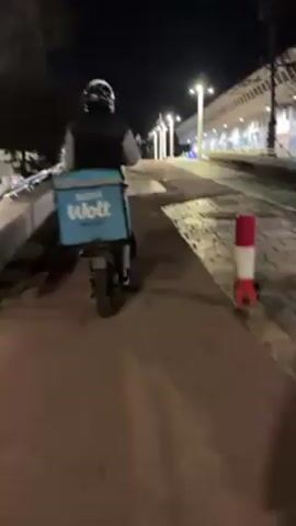 A person wearing a helmet and a Wolt delivery backpack rides an electric scooter on a pedestrian pathway at night. The pathway is paved with alternating sections of red and grey stone. Streetlights illuminate the scene. A large building with a white facade is visible on the right side of the pathway, and a few pedestrians walk in the distance on the left. A red and white striped traffic bollard stands near the right edge of the frame. The camera follows the scooter from behind as it moves forward.