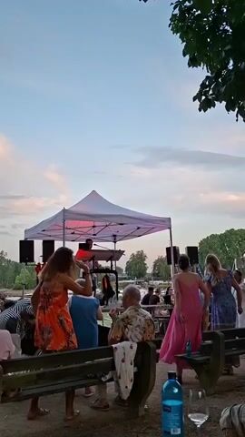 A live band performs under a white canopy during the day. A keyboard player is visible on stage, along with speakers. Several people are standing and dancing in front of the stage, while others are seated on benches. The setting appears to be outdoors, possibly near a body of water, with trees and a cloudy sky in the background.