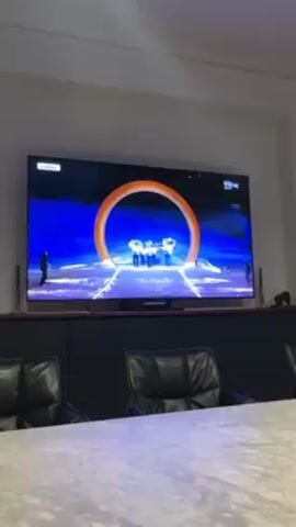 The video shows a television playing footage of the 2018 Winter Olympics closing ceremony. The ceremony shows athletes holding South Korean flags. The television is mounted on a dark gray cabinet with a concrete-looking conference table and black leather chairs in front of it. The wall is painted off-white.