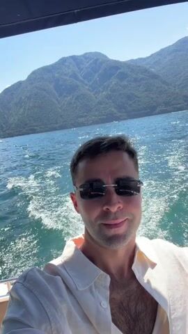 A man with short dark hair, wearing sunglasses, a light-colored open-collared shirt, and sunglasses, smiles while holding up a can of Birra Moretti. He is on a boat with blue water and mountains in the background. He takes a sip from the can.