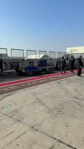 The video shows a pit lane at a racetrack. A blue and black race car is stationary in its pit. A group of people in dark clothing stand near the car, apparently involved in pit stop activities. One spare tire lies on the ground. The pit lane is marked with red lines. The track surface is concrete, showing tire marks. The sky is clear blue. A building with a green and white logo is visible in the background. The general atmosphere is that of a race event.