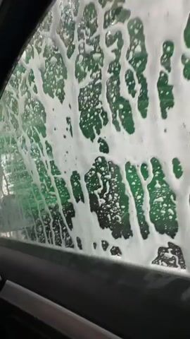 A first-person point-of-view video from inside a car driving through an automated car wash. The car is moving through various stages of the wash, with soap, water, and brushes visible on the windows and windshield. The car's interior is visible, including the dashboard and the driver's side door. The sun visor has a warning label in English and French about advanced air bags.