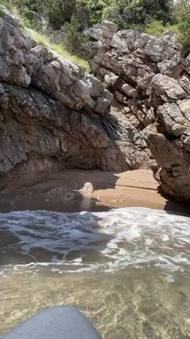 The camera is at water level, viewing a small cove between large grey rocks. The water is clear enough to see the sandy bottom. Small waves are gently washing onto the beach. The rocks are jagged and layered. Some greenery grows on top of the rock formations. The day is sunny, with strong light and shadows. A small cave can be seen in the rocks. There is a small patch of beach in the middle of the cove. The angle is slightly upwards, focusing on the rocks and beach.