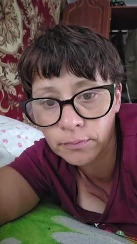 A woman with short, dark hair and glasses lies on her side, facing the camera. She is wearing a maroon shirt. She appears to be in a bedroom, with a patterned wall covering and a wooden cabinet visible in the background. The lighting is dim. The woman blinks and moves her head slightly, but otherwise remains still.