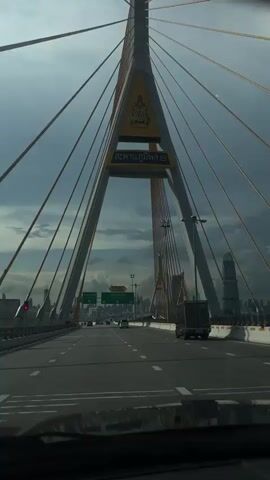 The video shows a view from inside a car driving on a cable-stayed bridge. The camera faces forward, looking down the road. A large yellow and white pylon with cables is visible in the center of the frame, with Thai text on it. Green road signs with white text are also visible, indicating directions and road names. The sky is cloudy and appears to be overcast, with sunlight filtering through in some areas. The cityscape of a city is visible in the distance. Other vehicles, including cars and a truck, are on the bridge. The video captures the movement of the car as it progresses across the bridge.
