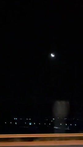 A crescent moon hangs in the dark night sky. The camera is in motion, possibly from a vehicle, as streaks of light and blurred shapes pass by. In the foreground, a faint, horizontal line suggests the edge of a road or structure. Distant lights twinkle on the horizon, indicating a town or city. The overall impression is one of traveling at night with the moon as a celestial guide.