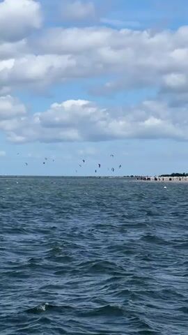 The video shows a wide shot of a body of water, likely the sea, with choppy waves under a partly cloudy sky. In the distance, along the horizon, a sandy beach is visible, lined with buildings and what appear to be beach umbrellas or tents. Scattered across the water, closer to the shore, are numerous kite surfers. They are identified by their colorful kites which are raised in the air. The kite surfers are engaged in their sport, some appearing to be in motion on the water. The overall scene depicts an active watersports location on a breezy day.