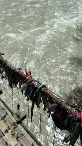 The video captures a scenic view from a suspension bridge over a powerful, fast-flowing river in Bzypta, Abkhazia.

The initial shot focuses on the weathered metal cables and wooden planks of the bridge's railing, which is adorned with numerous colorful ribbons and scraps of fabric, likely left by visitors as a tradition or wish. The turbulent, light-grey water of the river churns below, creating foamy white rapids.

As the camera pans upwards and to the right, it reveals the expansive river flowing through a valley. The riverbanks are lush with dense green trees and vegetation. In the background, majestic mountains rise, their slopes covered in vibrant green forests, extending towards a partially cloudy, bright sky. The scene depicts a serene yet powerful natural landscape, characteristic of a mountainous region. The lighting suggests it's daytime with good visibility. No people are visible in the video.