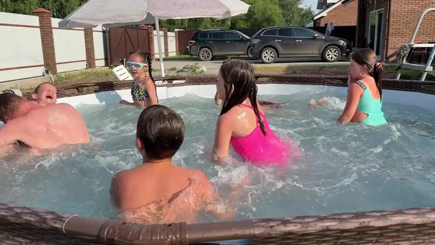 A group of children are playing in an above-ground swimming pool on a sunny day. A young boy with brown hair and sunglasses is in the foreground, facing away from the camera. To his left, an adult male with light brown hair is partially submerged in the water. To the boy's right, a girl with dark hair in a pink swimsuit is splashing water. Behind her, another girl with dark hair in a blue swimsuit is also splashing. In the background, two other children are visible in the pool. The pool is surrounded by a brown, woven material. A white umbrella is positioned over the pool. A brick fence and two parked cars are visible behind the pool. The sky is clear and the trees in the background are green.