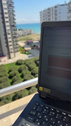 The video shows an Acer laptop on a balcony overlooking a cityscape and a body of water. The laptop screen displays a Google Sheets document in Russian titled "Plan-Fact Managers Ozon." The spreadsheet is filled with numerical data and labels related to sales, margin, and advertising expenses. In the background, there are tall apartment buildings, a green area with round bushes, a road, and a distant view of the sea under a clear blue sky. The camera pans across the laptop screen, showing different sections of the spreadsheet. The balcony railing is visible in the foreground. The laptop has an "HD 1080" sticker on it.