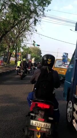 The video shows a street scene with multiple motorcycles on a paved road. In the foreground, a person wearing a black helmet and a backpack rides a red scooter with a license plate that reads L 2939 ABW. They are wearing blue pants. To the right, a blue truck is partially visible. Further ahead, several other motorcycles are also on the road, including one with a yellow sticker on its helmet. A yellow van is seen in the middle ground. A traffic light is visible in the distance, showing a green signal. Trees line the left side of the road. The sky is clear and appears to be daytime. The scene suggests traffic moving through an urban or suburban environment.