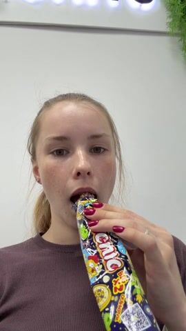 A young woman with light hair tied in a ponytail and fair skin with freckles is captured indoors in Usolye, Russia. She is wearing a dark purple long-sleeved top, and her fingernails are painted red. The central activity shows her eating from a large, colorful, tubular snack package. The package is adorned with cartoon characters and prominently features the words "GIG" and "BIG" in English, along with other foreign language text. She takes a bite from the package, chews, and then looks around with a neutral expression. The background consists of a plain white wall, a brightly lit white fluorescent sign with partially visible letters, and a small green artificial plant in the upper right corner, suggesting an indoor setting likely during the daytime.