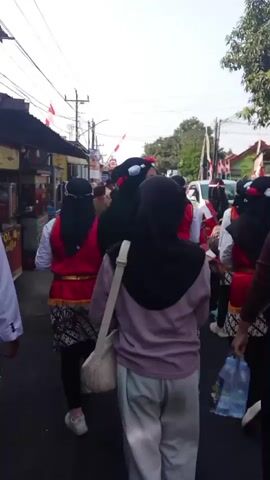 A group of people are walking down a street. Most of them are wearing traditional clothing, including red vests, black headscarves, and patterned skirts or trousers. Some individuals are carrying bags, including a light-colored shoulder bag and a clear plastic bag with water bottles. The street appears to be lined with buildings and trees, and there are electrical wires overhead. Red and white flags are visible, suggesting a celebration or event. The camera is following the group from behind.