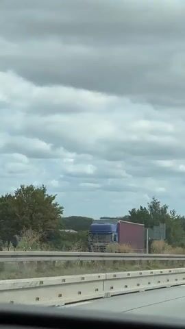 A video taken from a vehicle on a highway. The camera pans across the scene, showing other vehicles on the road, including cars and trucks. The environment is a highway with concrete barriers and a grassy shoulder. There are trees and fields visible in the background, with a cloudy sky overhead. The video appears to capture a normal highway scene with traffic.