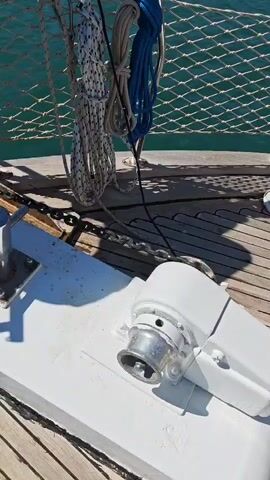 The video captures scenes aboard a boat, likely a sailboat, on a bright and clear day in Demre, Turkey.

The initial close-up shot focuses on the boat's foredeck, showing a white electric windlass (winch) actively operating, characterized by a distinct mechanical sound. A metal anchor chain feeds into the windlass's gypsy. Above the deck, various coiled ropes, including white and blue ones, are neatly stowed and hang from a metal railing. Beyond the railing, the clear, turquoise-blue water of the sea is visible.

The second shot pans to show a person, dressed in a dark t-shirt, black pants, and grey sneakers with yellow soles, standing on the boat's white deck. The person is bent forward, holding onto a polished metal railing, appearing to oversee the anchor operation or attend to something near the bow. The sound of the windlass continues.

The final shot presents a panoramic view from the boat, looking out over the water. The expansive sea is a vibrant blue-green, sparkling with sunlight reflections on its calm surface. In the distance, rolling green hills and mountains line the horizon, characteristic of a coastal landscape. Several other boats or yachts can be seen anchored or cruising in the serene bay. The sky is a clear, unblemished blue, indicating excellent weather.

The primary activity depicted is the operation of the boat's anchor system, with the windlass audibly working throughout the clips. The overall scene conveys a peaceful and sunny day on the water in a scenic coastal region. No visible text is present in the video.