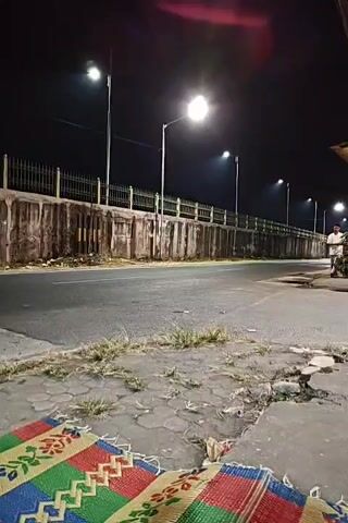 A night scene on a paved road under streetlights. A person on a motorcycle rides past.